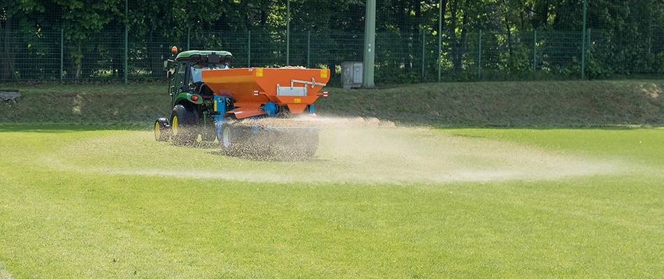 A tractor dispersing granular fertilizer over an athletic field to improve the lawn's health.